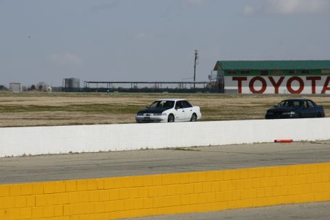 1995 Ford Taurus - A white sedan with a black hood is on a racetrack, with another dark car in the background and a Toyota building.