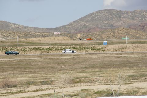 1995 Ford Taurus - Two cars are driving on a racetrack with hills and a blue portable toilet in the background.