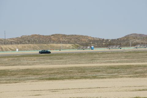 1995 Ford Taurus - A lone dark sedan drives on a paved track with dry hills and empty bleachers under a hazy sky.