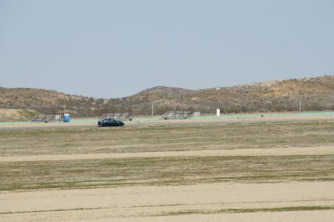 1995 Ford Taurus - A dark sedan drives on a paved track with dry grass and hills under a hazy sky.