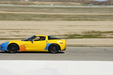 1995 Ford Taurus - A yellow sports car with custom blue and orange paintwork speeds along a race track under a clear sky.