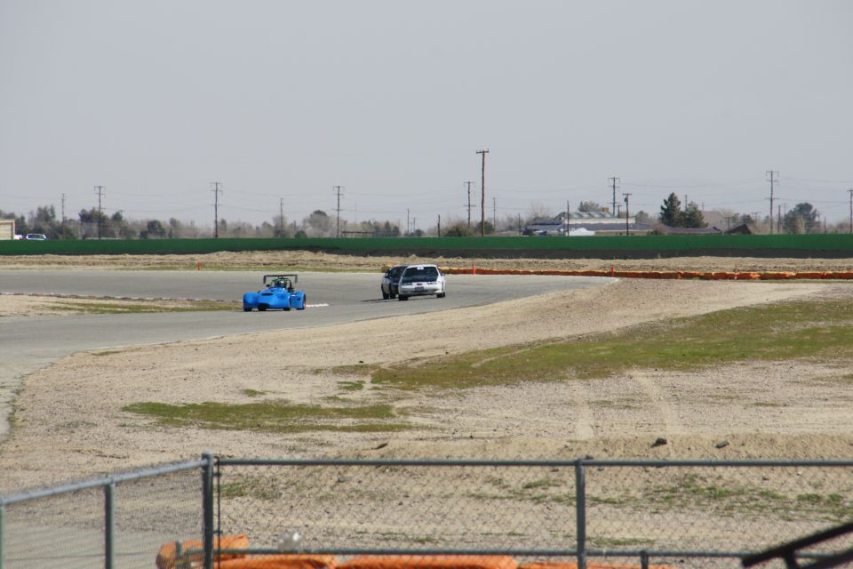 Blue race car and silver sedan on a racetrack with a chain-link fence in the foreground