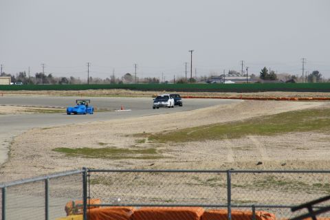 1995 Ford Taurus - A blue race car and two sedans are on a race track with a fence in the foreground.