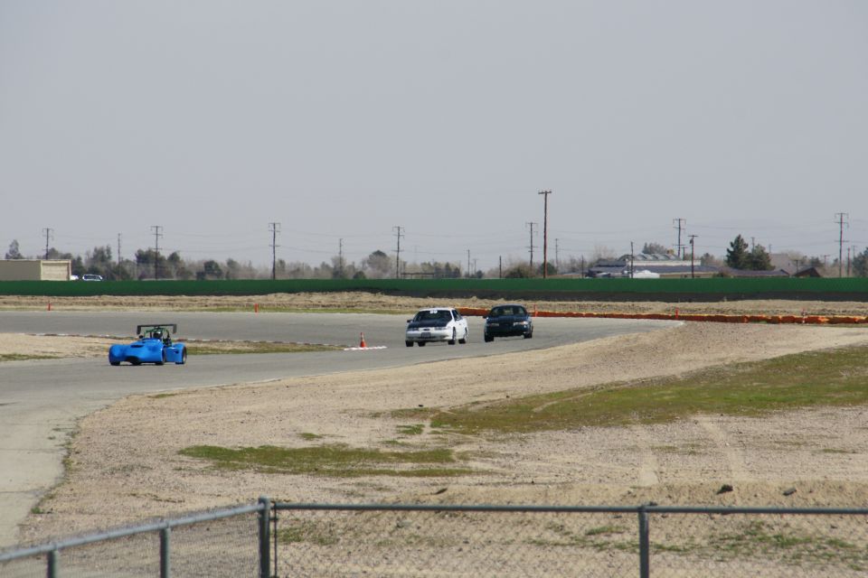 Blue sports car and two sedans on a race track, with a fence in the foreground