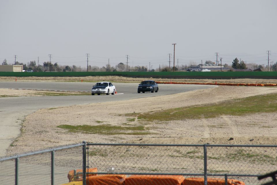 Two sedans race on a track, one white with black hood, one dark