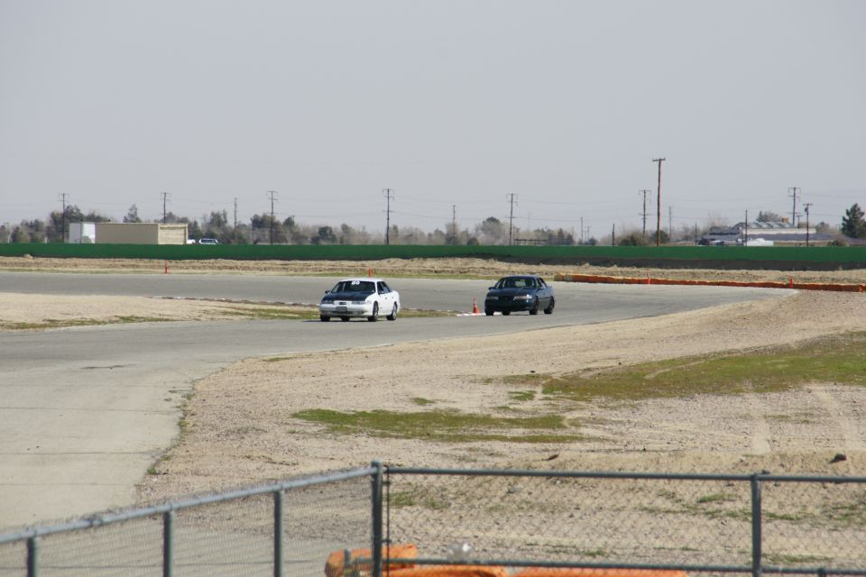 Two sedans, one white with black hood and one dark, race on a track with orange cones