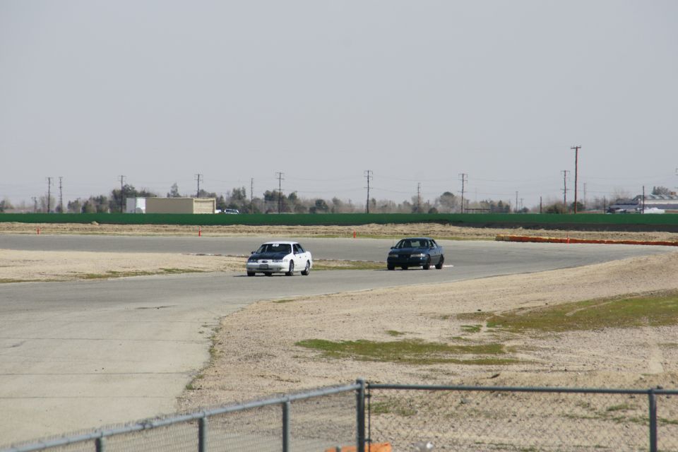 Two cars race on a track, one white with black hood, one dark colored