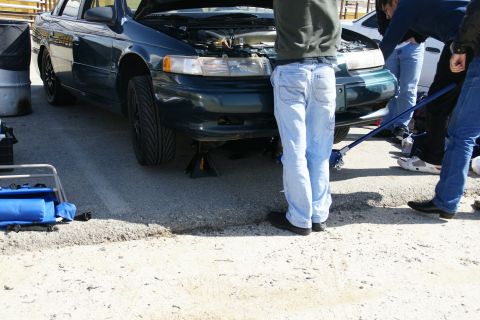 1995 Ford Taurus - Several people are gathered around a dark green sedan that is raised on jack stands, with its hood open.