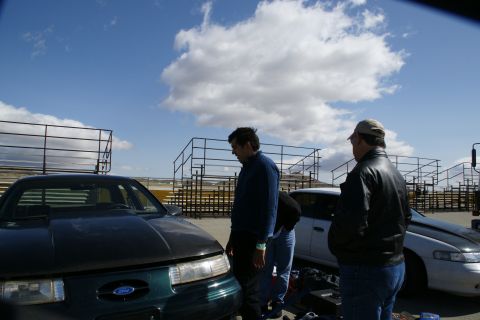 1995 Ford Taurus - A dark green 1995 Ford Taurus with an aftermarket hood is parked at a track with two men standing nearby.