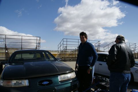 1995 Ford Taurus - Two men stand by a dark green 1995 Ford Taurus at an outdoor event with bleachers in the background.