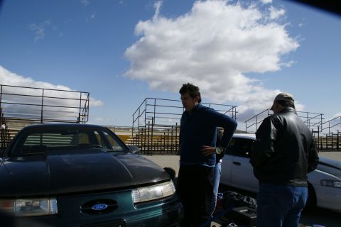1995 Ford Taurus - Two men stand near a dark green 1995 Ford Taurus with an aftermarket hood at an outdoor venue.