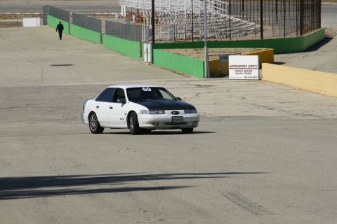 1995 Ford Taurus - A white 1995 Ford Taurus with a black hood drives on a race track, with a person walking in the background.
