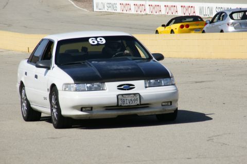1995 Ford Taurus - A white 1995 Ford Taurus with a black hood and racing number 69 is seen on a race track, with other vehicles in the background.