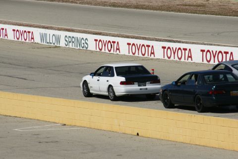 1995 Ford Taurus - Two sedans, one white and one dark, race on a track with 'TOYOTA' and 'WILLOW SPRINGS' signage.