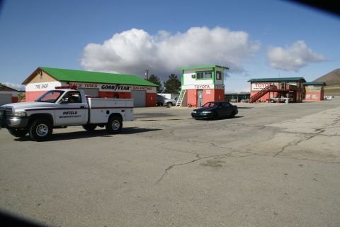 1995 Ford Taurus - A service truck and a sedan are parked on the asphalt at a racetrack with various buildings in the background.