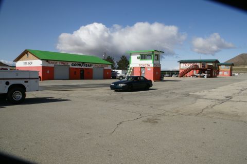 1995 Ford Taurus - A dark sedan drives past buildings at a race track, including a tire shop and a Toyota dealership.