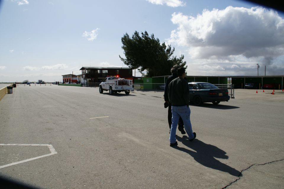 Two people stand on a paved lot near a utility truck with flashing lights and a dark sedan
