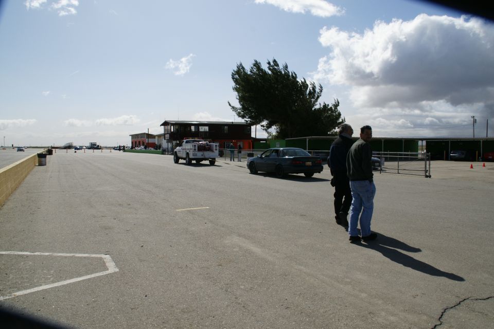 people standing near a dark sedan and a white pickup truck on a paved area with buildings and a tree in the background
