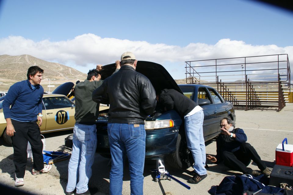 Several people are gathered around the open hood of a dark sedan, appearing to work on it. Another car with the number 07 is parked nearby