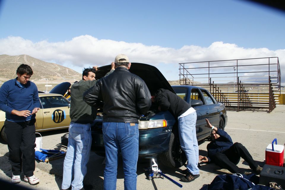 Dark green sedan with hood open, jacked up, with people working on it. Yellow car with '07' visible behind it
