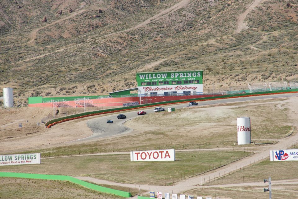 Several cars race on a track with Willow Springs sign and Budweiser Balcony in background