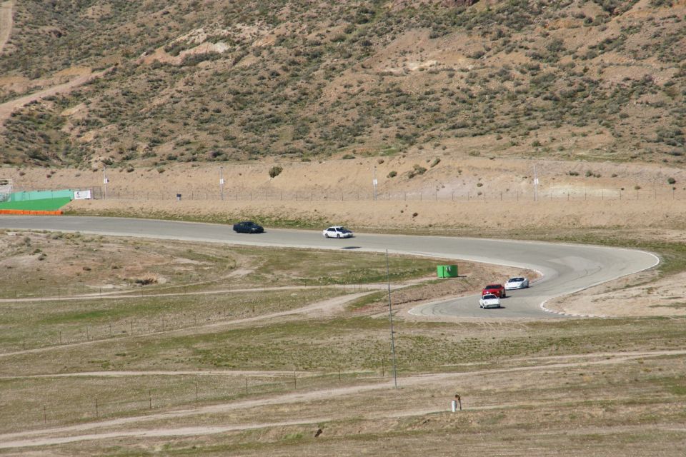 Several cars drive on a winding asphalt racetrack in a dry, hilly landscape