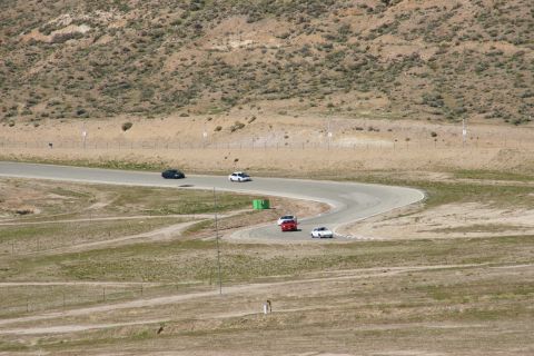 1995 Ford Taurus - Multiple vehicles navigate a turn on a paved racetrack surrounded by arid terrain.