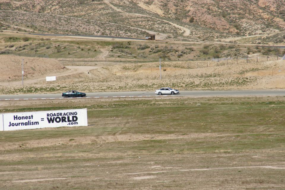 White sedan and dark coupe race cars on a track with a "ROADRACING WORLD" sign