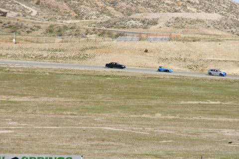 1995 Ford Taurus - A black sedan, a blue race car, and a silver sedan drive on a racetrack with dry, hilly terrain in the background.