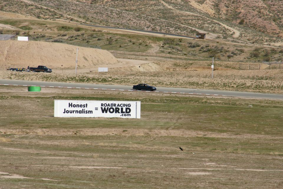 Black sedan driving on a racetrack with a truck and trailer parked nearby. Sign says "Honest Journalism = ROADRACING WORLD .com"