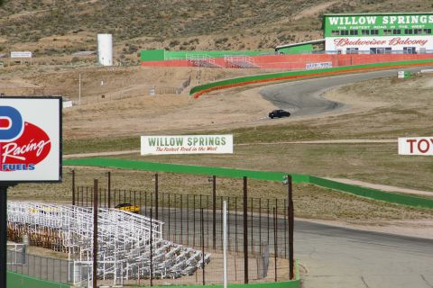 1995 Ford Taurus - A black sedan navigates a turn on the Willow Springs race track, with a yellow vehicle visible near the bleachers.