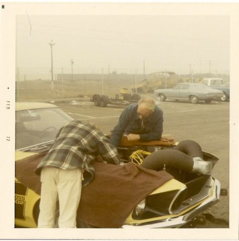 1973 Ford Mustang - Two men are working on the engine of a yellow muscle car, with other vehicles in the background.