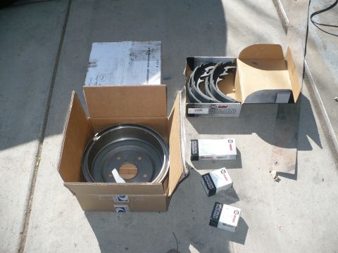 1979 Ford Bronco - New brake parts laid out on a concrete driveway: a bare five-lug brake drum sitting in its open cardboard box, a boxed set of new brake sho…