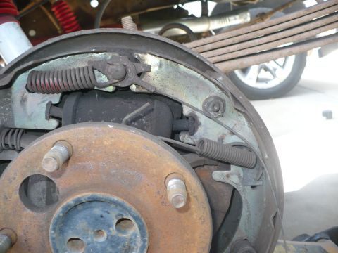 1979 Ford Bronco - Tight close-up of a worn rear drum brake with the drum off, showing dark used shoes, red and bare return springs, the adjuster lever and a…