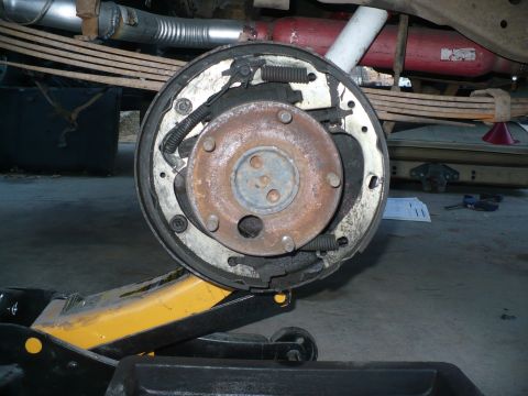 1979 Ford Bronco - Rear drum brake with the drum pulled off, showing worn shoes, return springs and a rusty five-lug hub caked with brake dust on a white-pain…