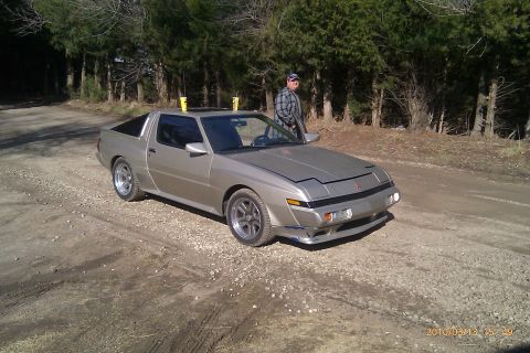 2000 Mercury Grand Marquis - A man stands next to a silver Mitsubishi Starion on a gravel road with trees in the background.