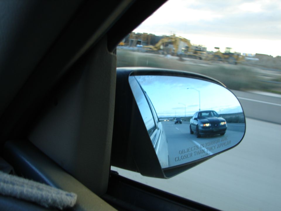 Car in side mirror on highway, construction in background. Text on mirror reads 'OBJECTS IN MIRROR ARE CLOSER THAN THEY APPEAR'