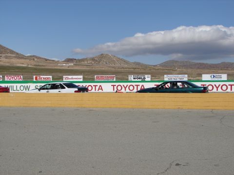 1992 Ford Taurus - Two sedans, a white one and a dark green one, are positioned side-by-side on a race track with mountains in the background.