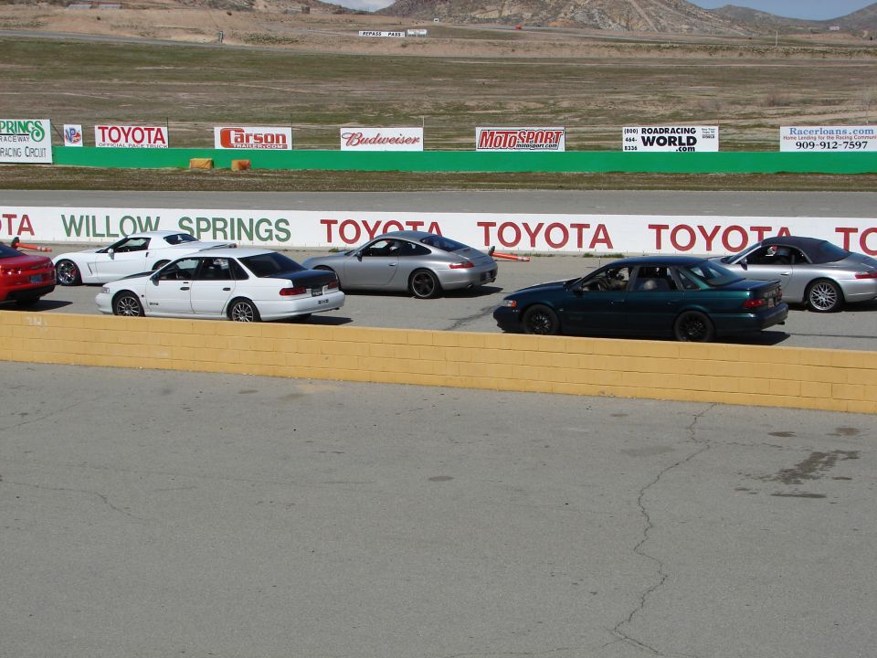 Cars on a race track, including a white sedan with a black trunk and a dark green sedan