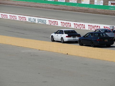 1992 Ford Taurus - A white 1992 Ford Taurus with a black trunk leads a dark sedan on a race track with Toyota branding on the wall.