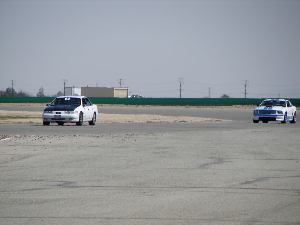 Two cars race on a track, one with a black hood and the other with blue stripes