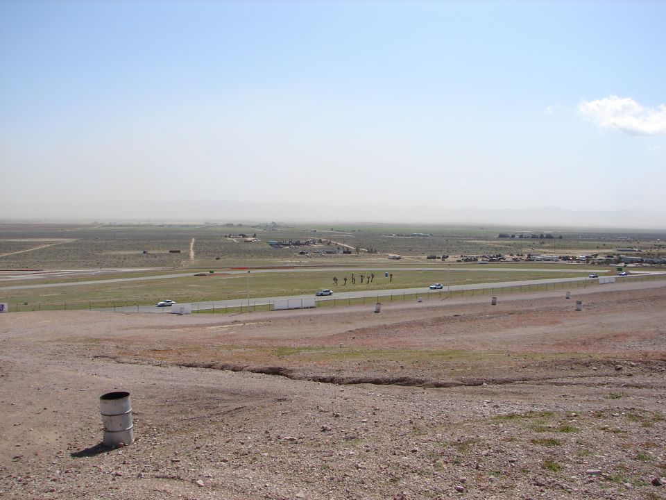 Race track scene with distant buildings and mountains under a hazy sky. A few cars are visible on the track