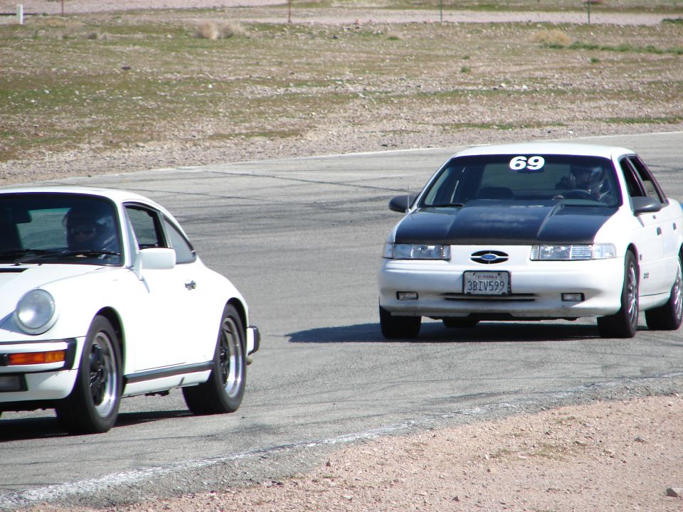 White 1992 Ford Taurus with black hood and number 69 on windshield races a white Porsche on a track