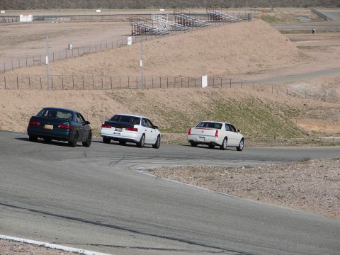 1992 Ford Taurus - Three sedans are captured from behind as they navigate a turn on a paved race track with grandstands in the background.