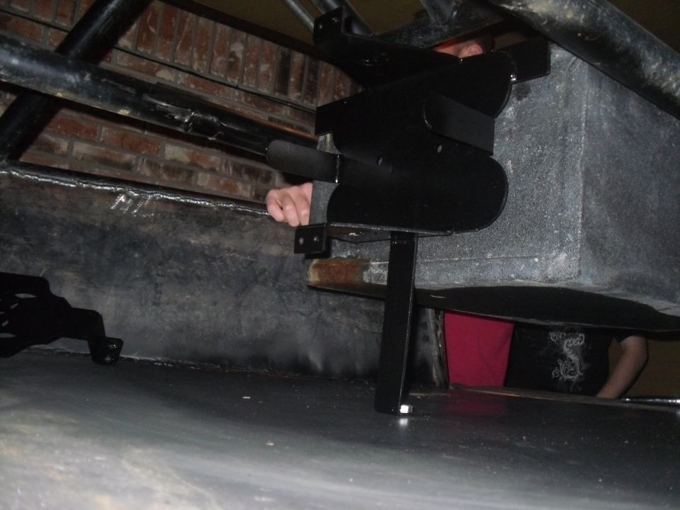 Underside view of a black metal bracket attached to a grey rectangular component, with a person's arm visible