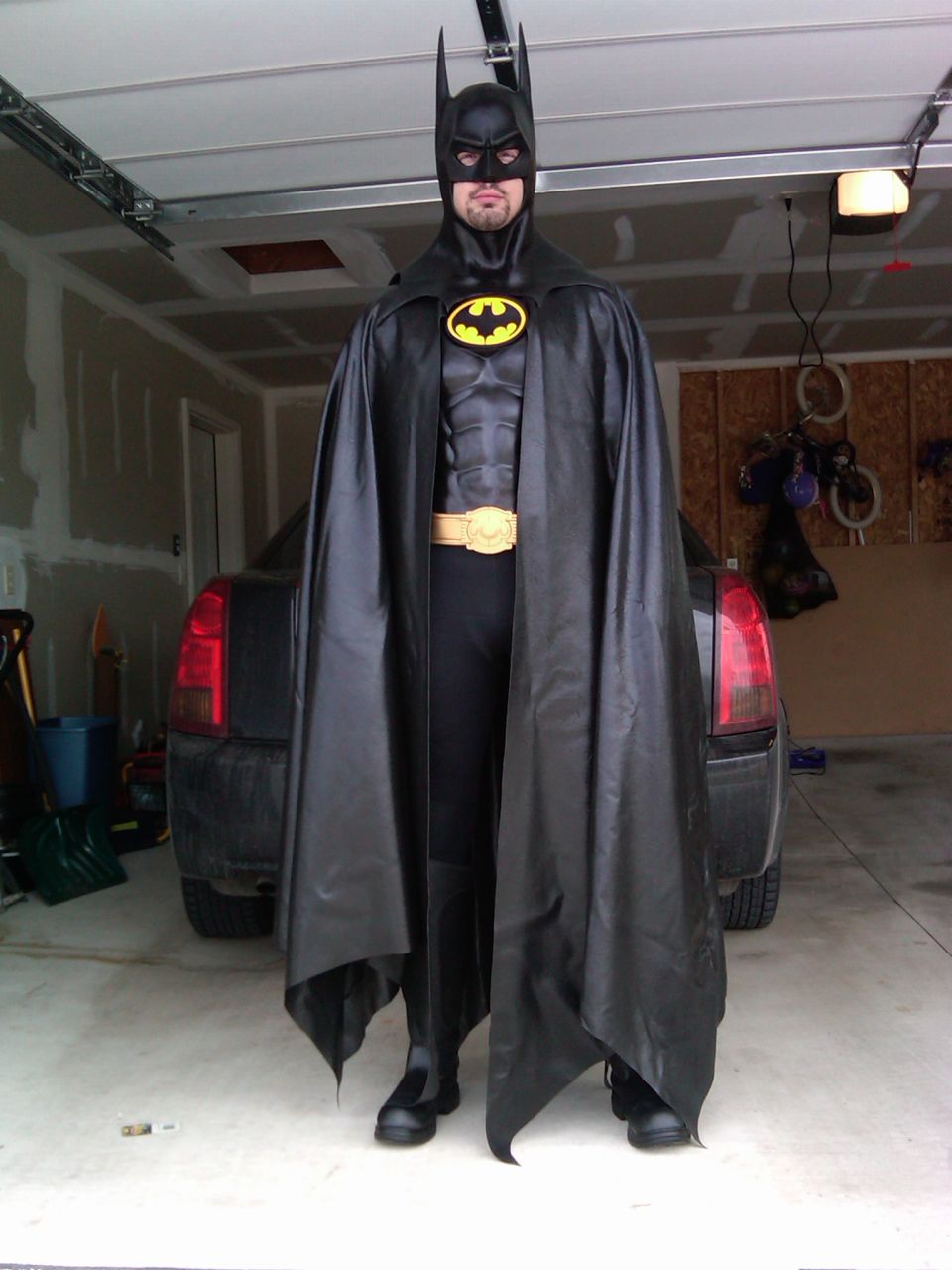 Man in black Batman costume with yellow belt and bat symbol standing in a garage