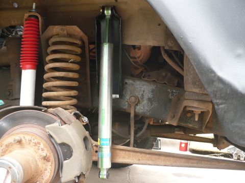 1979 Ford Bronco - Close-up of the front suspension and brake assembly on a lifted truck with the wheel removed, showing a rusty coil spring, a red-boot white…