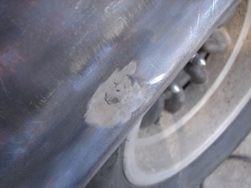 Close-up of a car's metal body panel showing a patch of body filler over rust damage, with a tire and wheel visible in the background