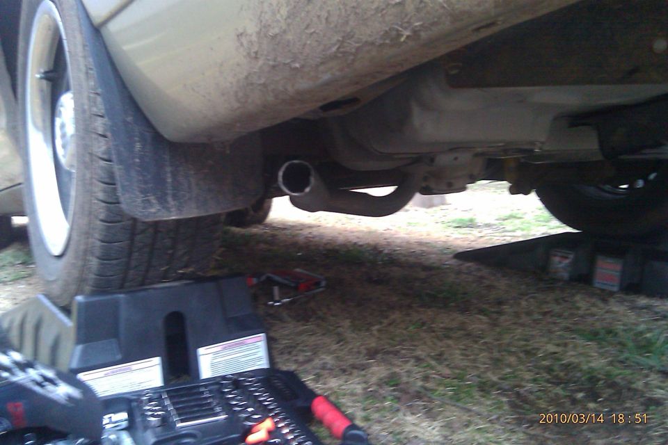 Underside of a light-colored car, showing the exhaust pipe and rear tire on a jack stand