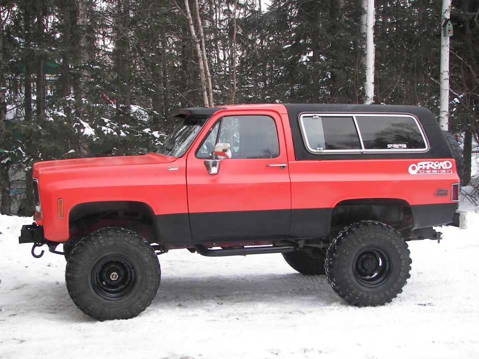 Red and black lifted SUV with off-road tires, side view, snow on ground, trees in background